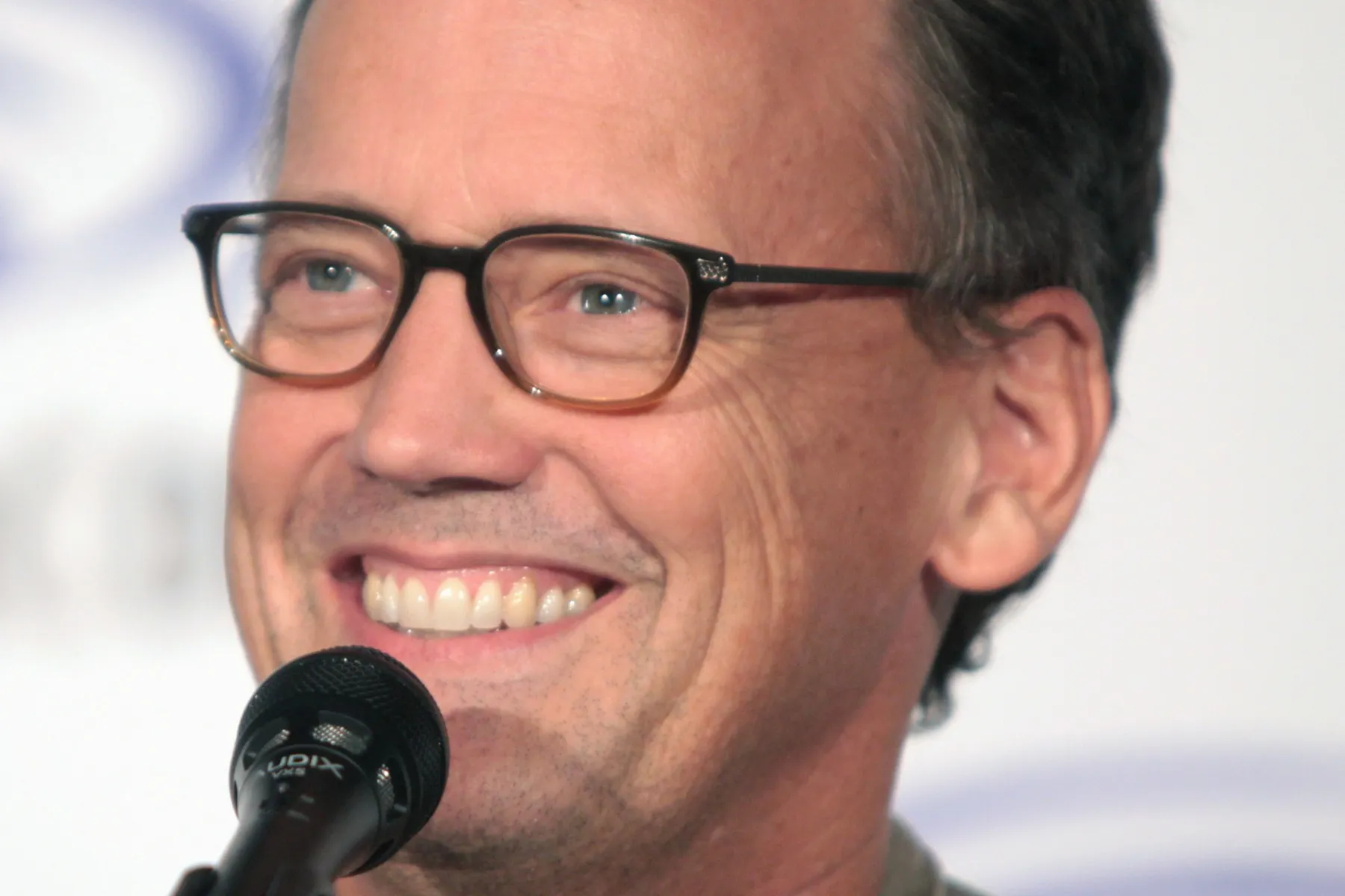 Dee Bradley Baker smiling and speaking into a microphone, wearing glasses.